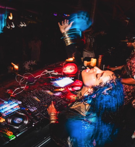 A BAE DJ with blue hair under red light, arms raised