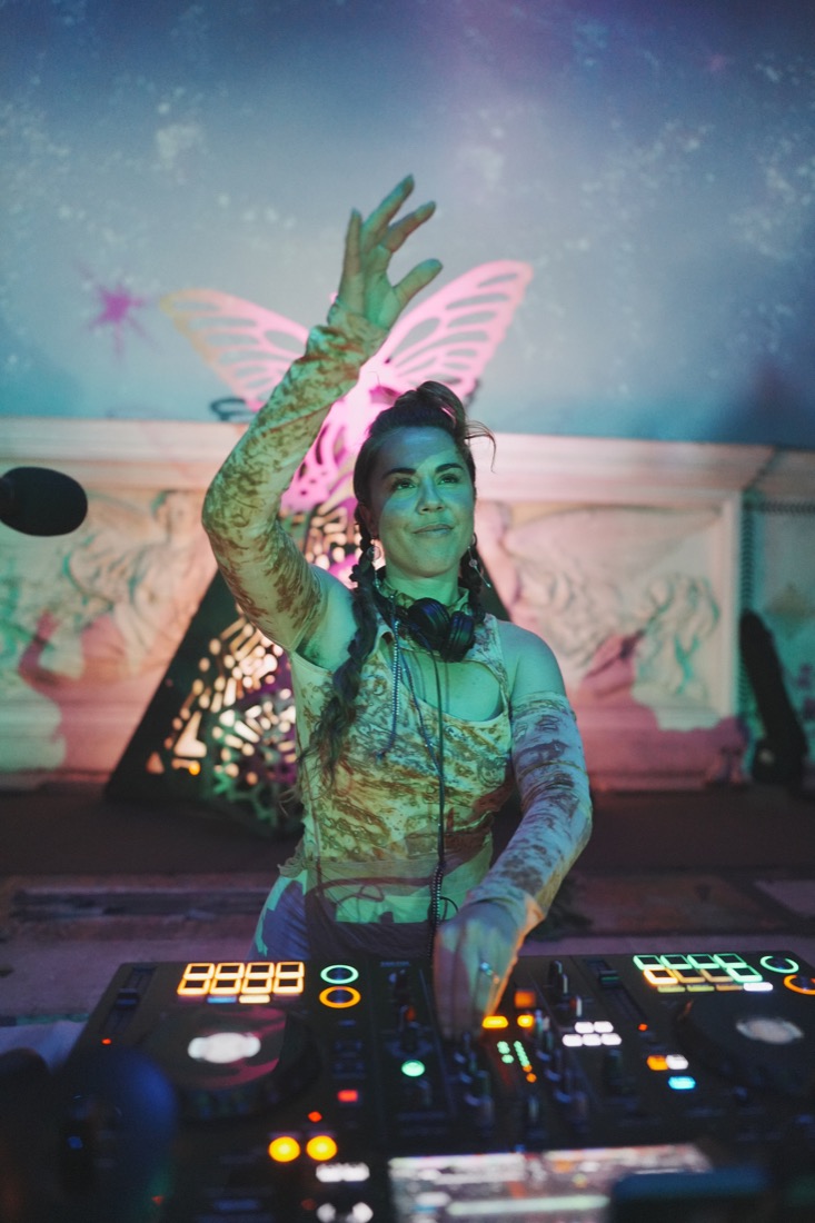 A BAE DJ with a raised hand before glowing pink butterfly wings